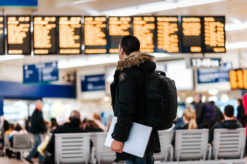 man holding laptop in an airport while looking at the flight departure board